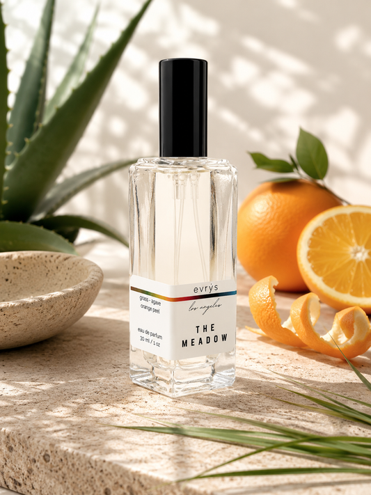Perfume bottle labeled 'The Meadow' on a stone surface with oranges and a plant.