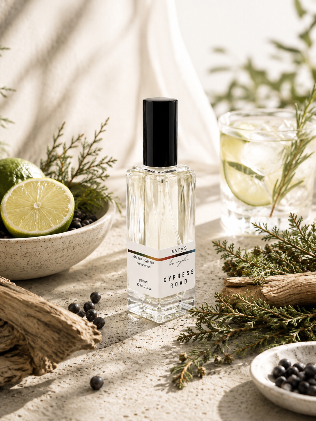Perfume bottle labeled 'Cypress Road' surrounded by limes, herbs, and a gin and tonic on a textured surface.