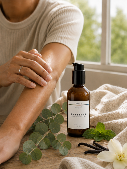 Person applying DAYBREAK lotion to their arm with elements of vanilla and eucalyptus on a wooden surface.