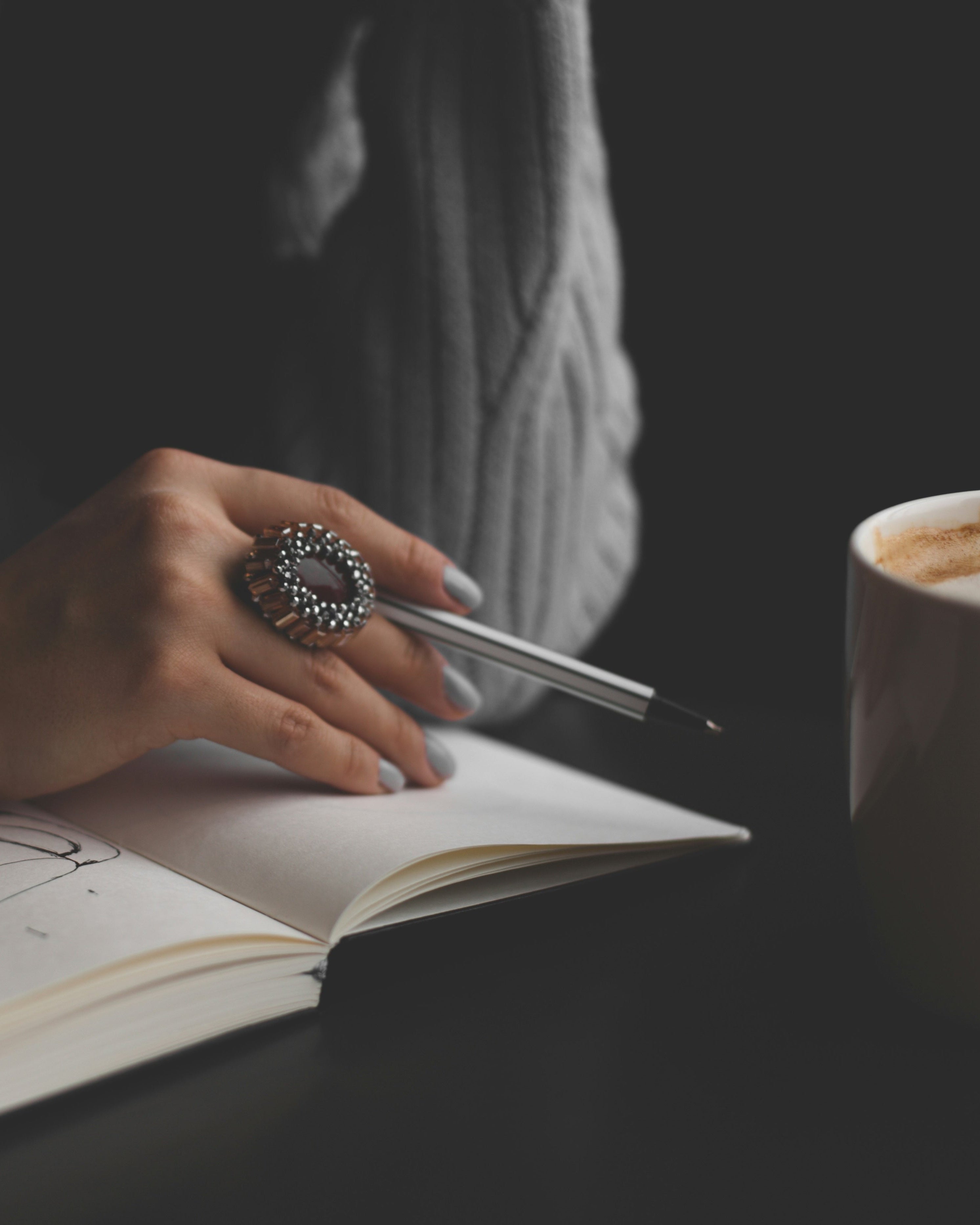 Person writing in a notebook with a pen next to a cup of coffee on a dark surface