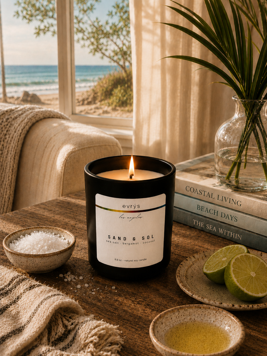 Candle labeled 'Sand & Sol' on a table with a coastal view through a window.