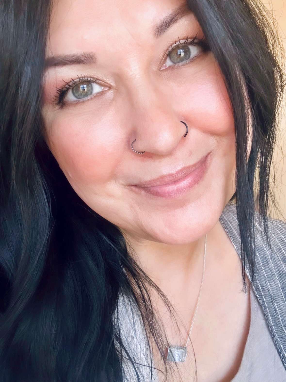 Close-up of a person smiling with dark hair and nose rings, wearing a necklace.